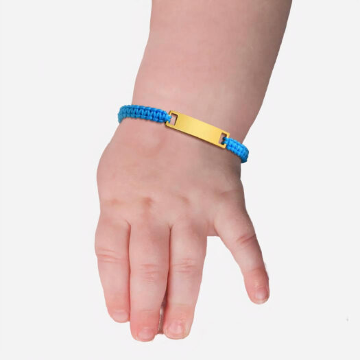 L'avant bras d'un enfant porte un bracelet avec un cordon bleu et une plaque dorée. Il porte un habit en dentelle blanche