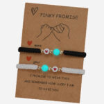 Deux bracelets amitié noire et blanc avec une perle phosphorescente et une perle avec un coeur, tout les deux sur un présentoir en carton.