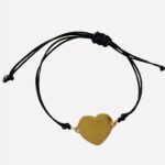 Un bracelet avec un cordon noir fermé par un noeud et un coeur doré en breloque. Il est posé sur un tissu moumoute blanc