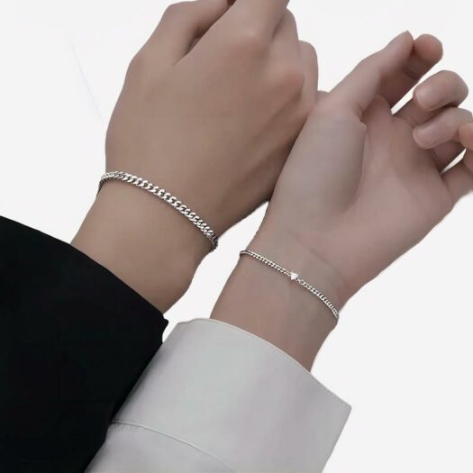 Photo de mains d'un couple portant des bracelets assortis aux poignets en mailles gormette de taile différente avec un petit coeur en cristal sur le modèle de la femme, les bras sont posés sur un support beige et les mains sur un papier blanc