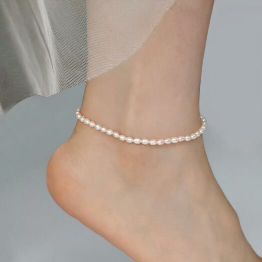 Photo d'une cheville de femme portant un bracelet en perles d'eau douce de forme grain de riz blanc sur un fond gris