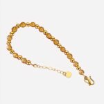 Un bracelet en perles dorées ouvert. Petite chaine d'ajustement avec un coeur. Le bracelet est posé sur un tissu rouge