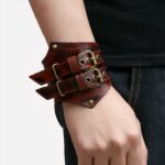 Photo de l'avant-bras d'in homme portant un bracelet de force rouge en cuir à deux boucles au look rock et deux rivets au bout des pointes de devant, l'homme porte des vêtements noirs et le fond de la photo à gauche est beige