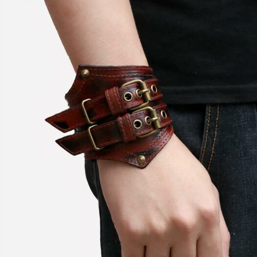 Photo de l'avant-bras d'in homme portant un bracelet de force rouge en cuir à deux boucles au look rock et deux rivets au bout des pointes de devant, l'homme porte des vêtements noirs et le fond de la photo à gauche est beige