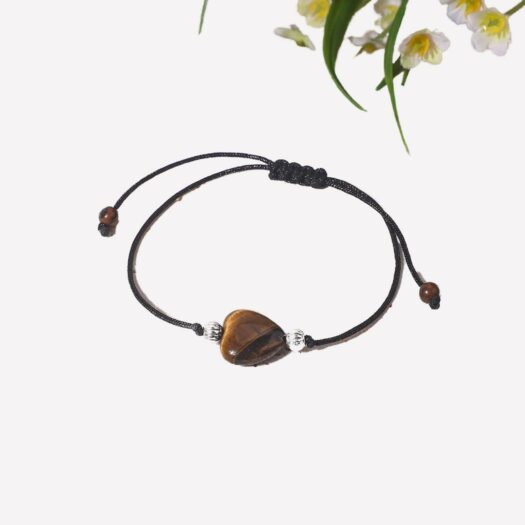 Photo d'un bracelet oeil de tigre en forme de coeur pour fille et adolescente sur un cordon de coton et deux perles argentées qui entourent le coeur, bracelet sur une pierre rectangulaire en grès beige avec une fleur en haut de la photo et du rose saumon autour de la pierre