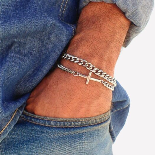 Un homme porte au poignet 2 barcelets chaines argenté. Un des bracelets a une croix. La main est dans la poche d'un jean
