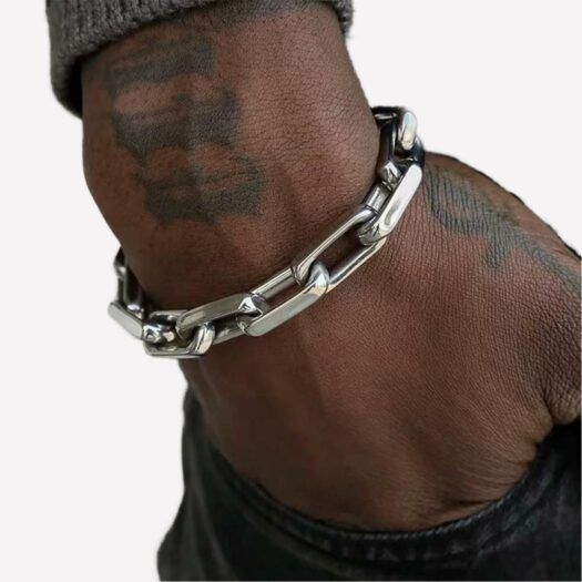 Un poignet porte un bracelet en métal gris avec de grosses mailles. La main ets dans une poche
